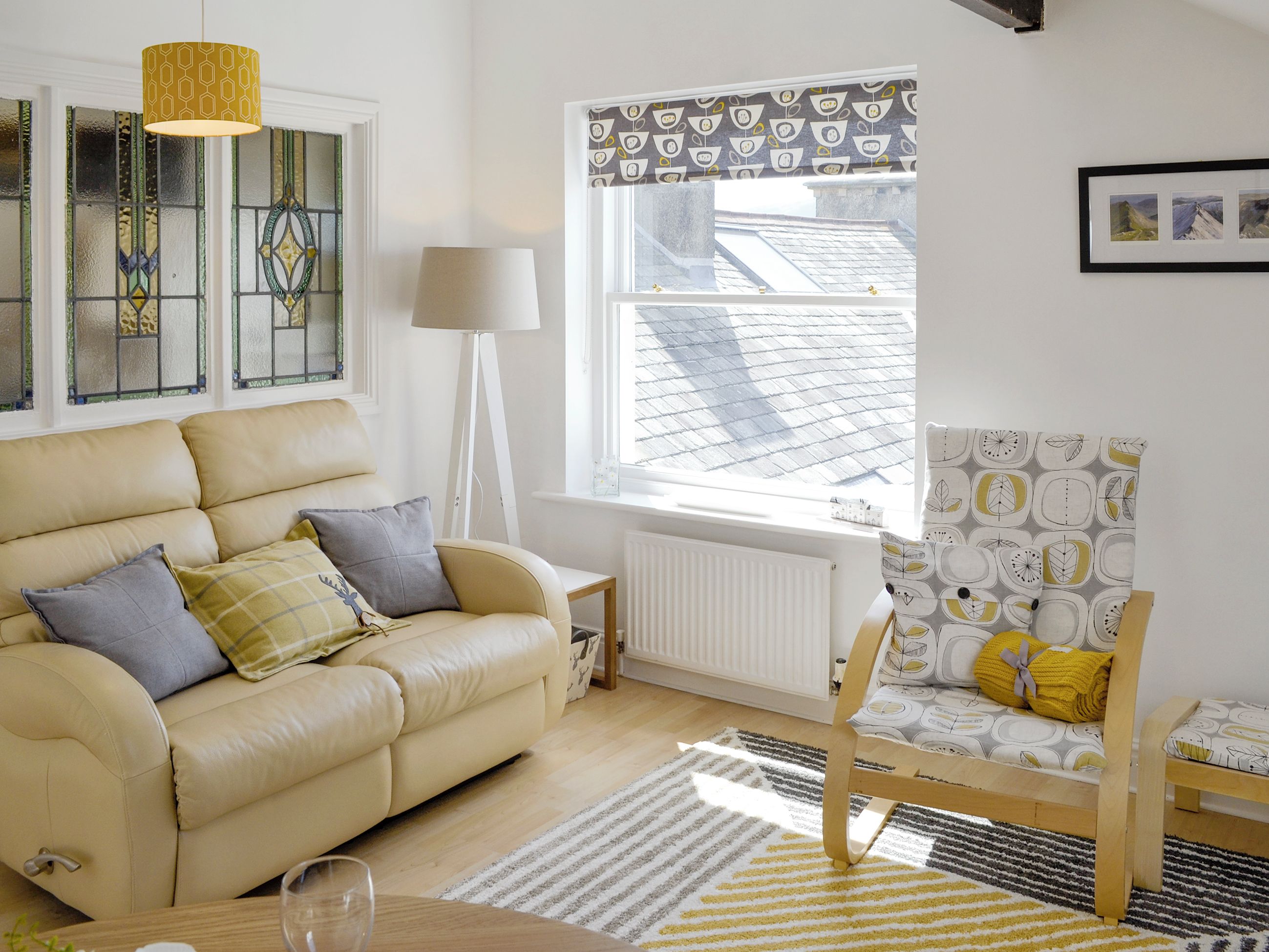 A cozy living room with a beige sofa, a wooden chair, a floor lamp, and a view of the roof through a window.