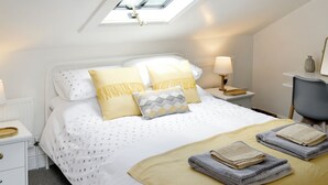 Ein Schlafzimmer mit Oberlicht, einem Bett mit gemusterten Betttextilien, Nachttischlampen, einem Sessel und einem kleinen Tisch mit einer Vase.