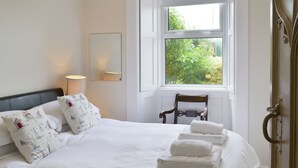 A neatly made bed with white linens and decorative pillows, a bedside lamp, a chair, and a window with a view of greenery. - 3 bedroom accommodation in Alnmouth, Alnwick (Alnmouth, Alnwick)