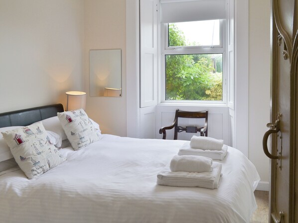 Una cama impecablemente arreglada con sábanas blancas y almohadas decorativas, una lámpara de noche, una silla y una ventana con vistas a zonas verdes.