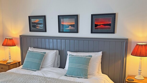 Een bed met witte lakens en blauwe kussens, twee nachtlampjes, en drie ingelijste foto’s aan de muur.