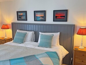 Een bed met witte lakens en blauwe kussens, twee nachtlampjes, en drie ingelijste foto’s aan de muur.