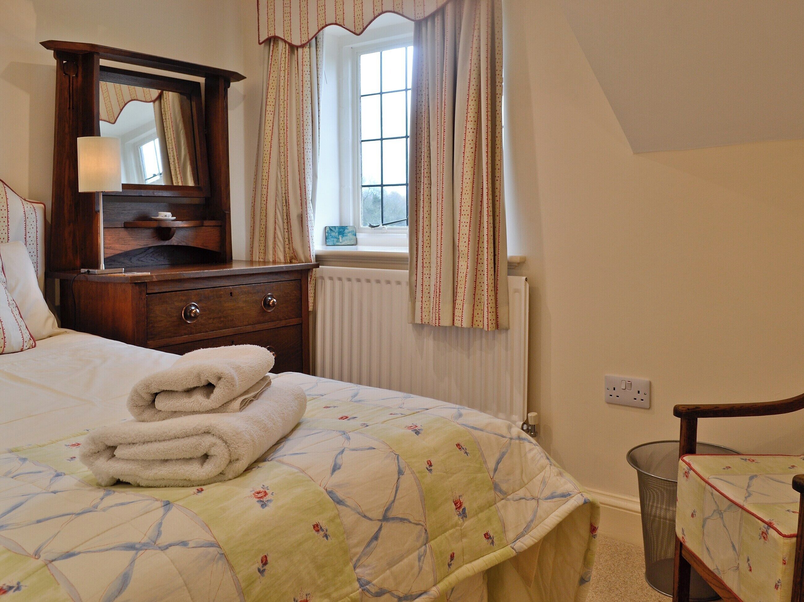 A neatly made bed with a floral quilt, a wooden dresser with a mirror, a window with curtains, and a radiator.