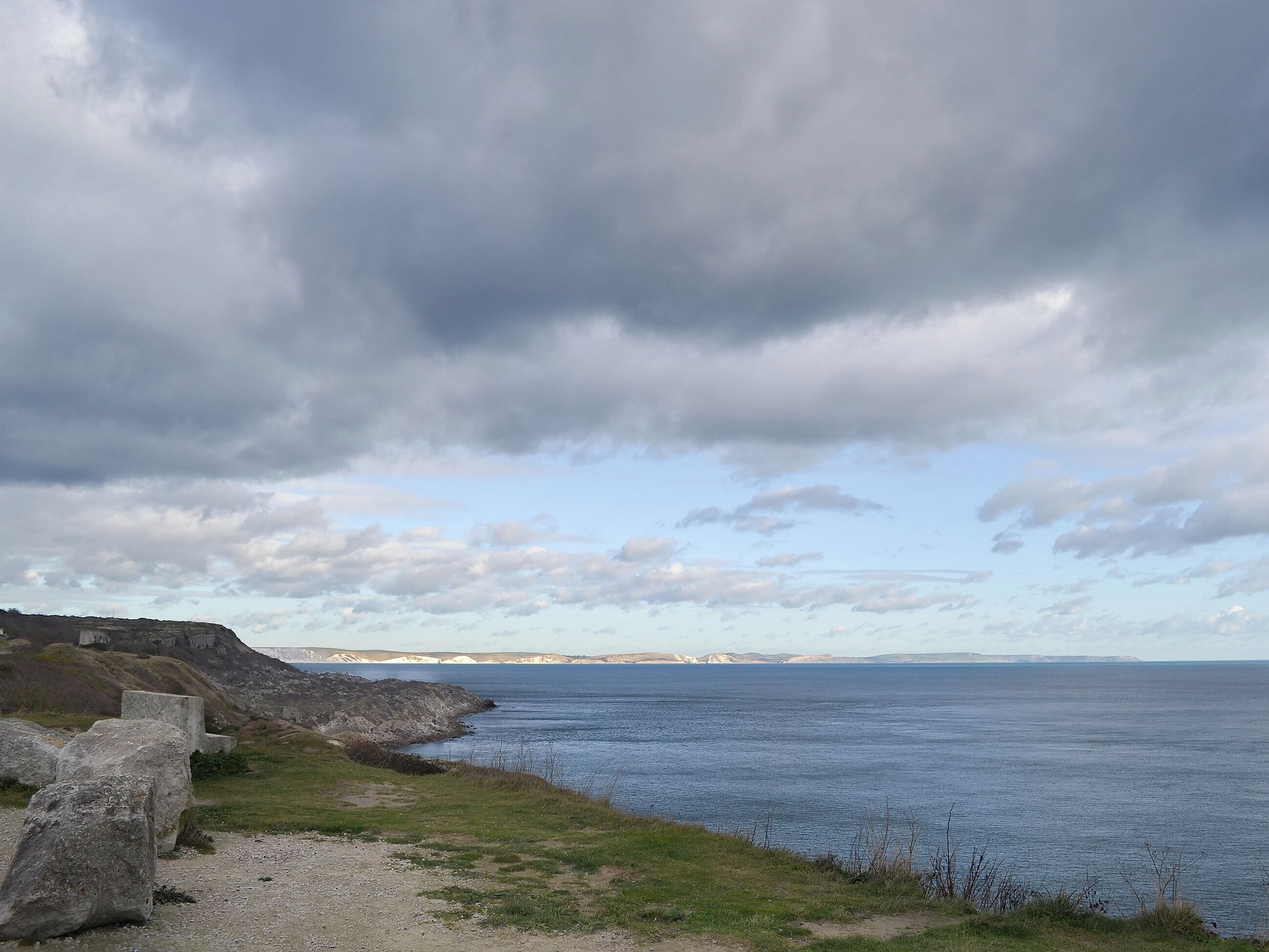 Paisaje costero con costa rocosa, vista despejada del mar y formaciones terrestres distantes bajo un cielo nublado.