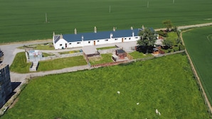 A white building with a blue roof, surrounded by green fields and a body of water in the background. - 3 bedroom accommodation in Portmahomack, near Tain (Portmahomack, near Tain)