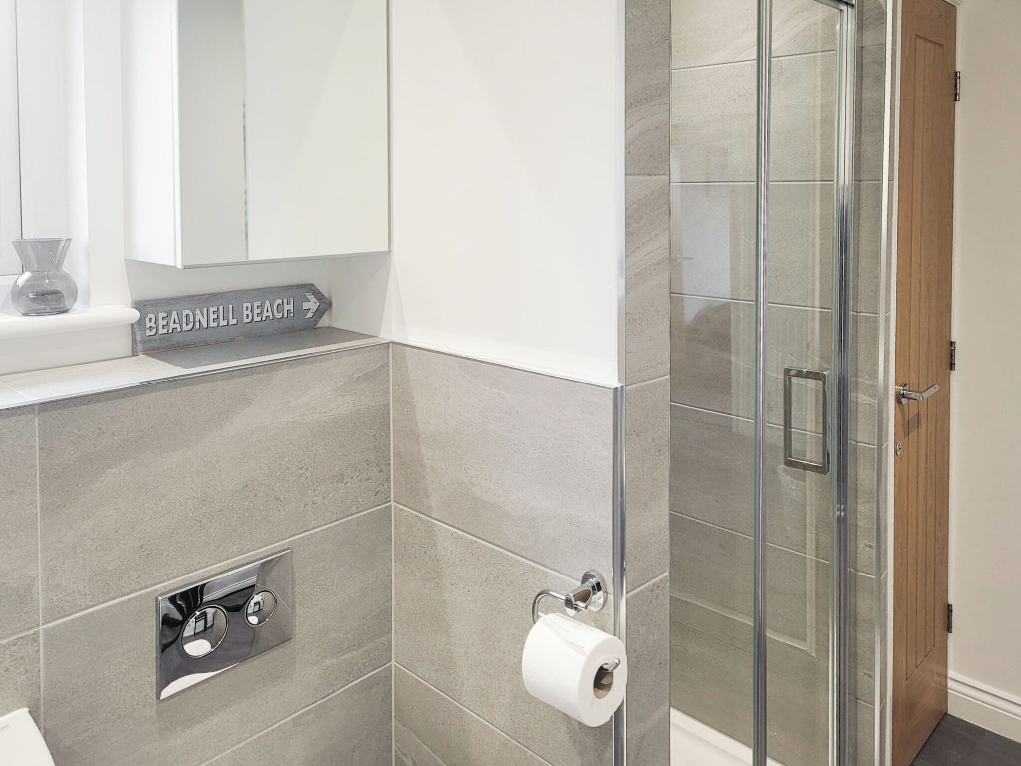 Une salle de bain avec des carreaux gris, un porte-rouleau de papier toilette, une douche avec une porte en verre et un panneau sur le mur.