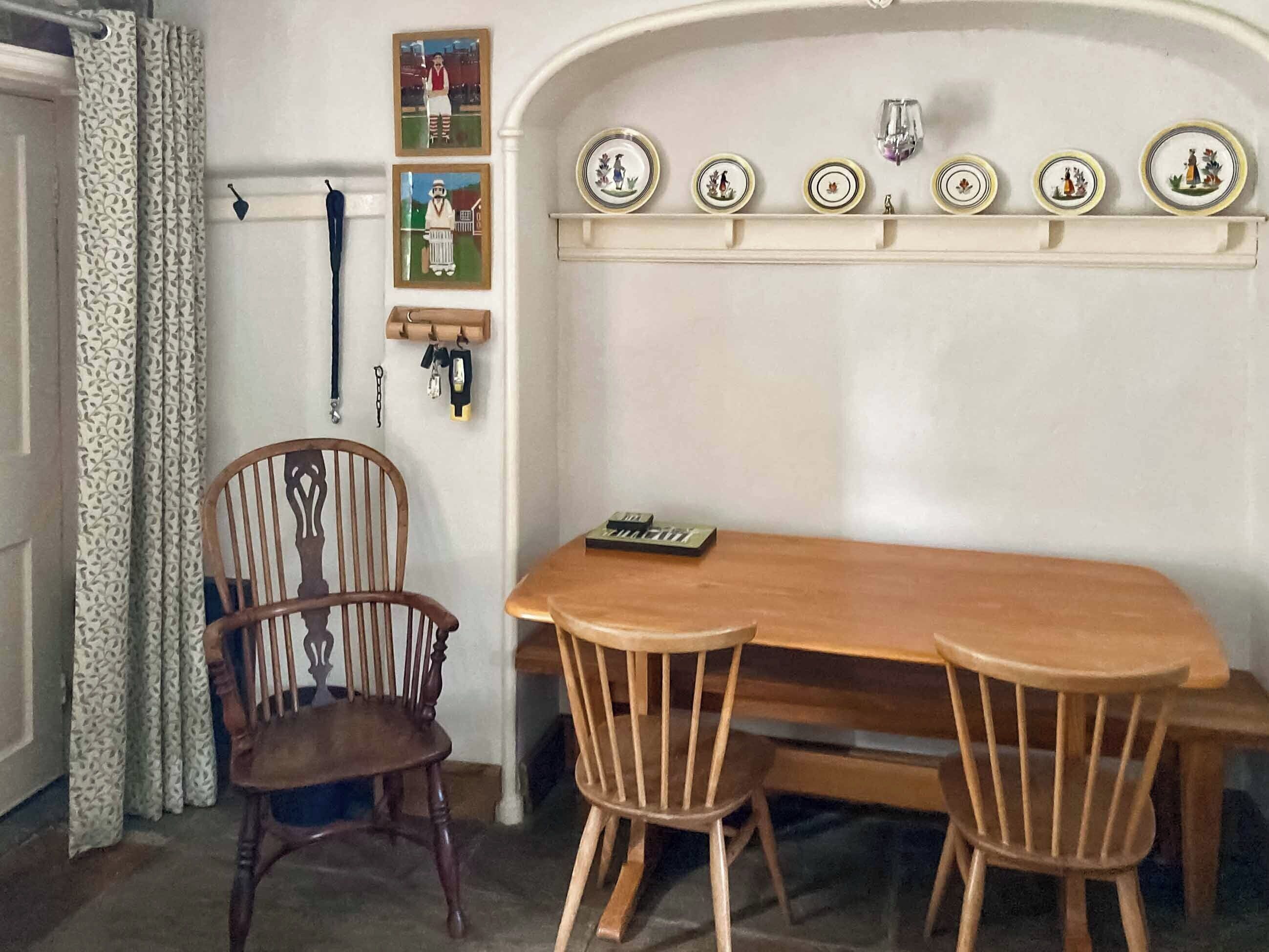 Un comedor con una mesa de madera y dos sillas, un perchero con llaves, y una estantería con platos decorativos.