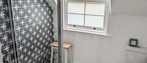 Un baño con ducha, una ventana, inodoro y un taburete de madera.