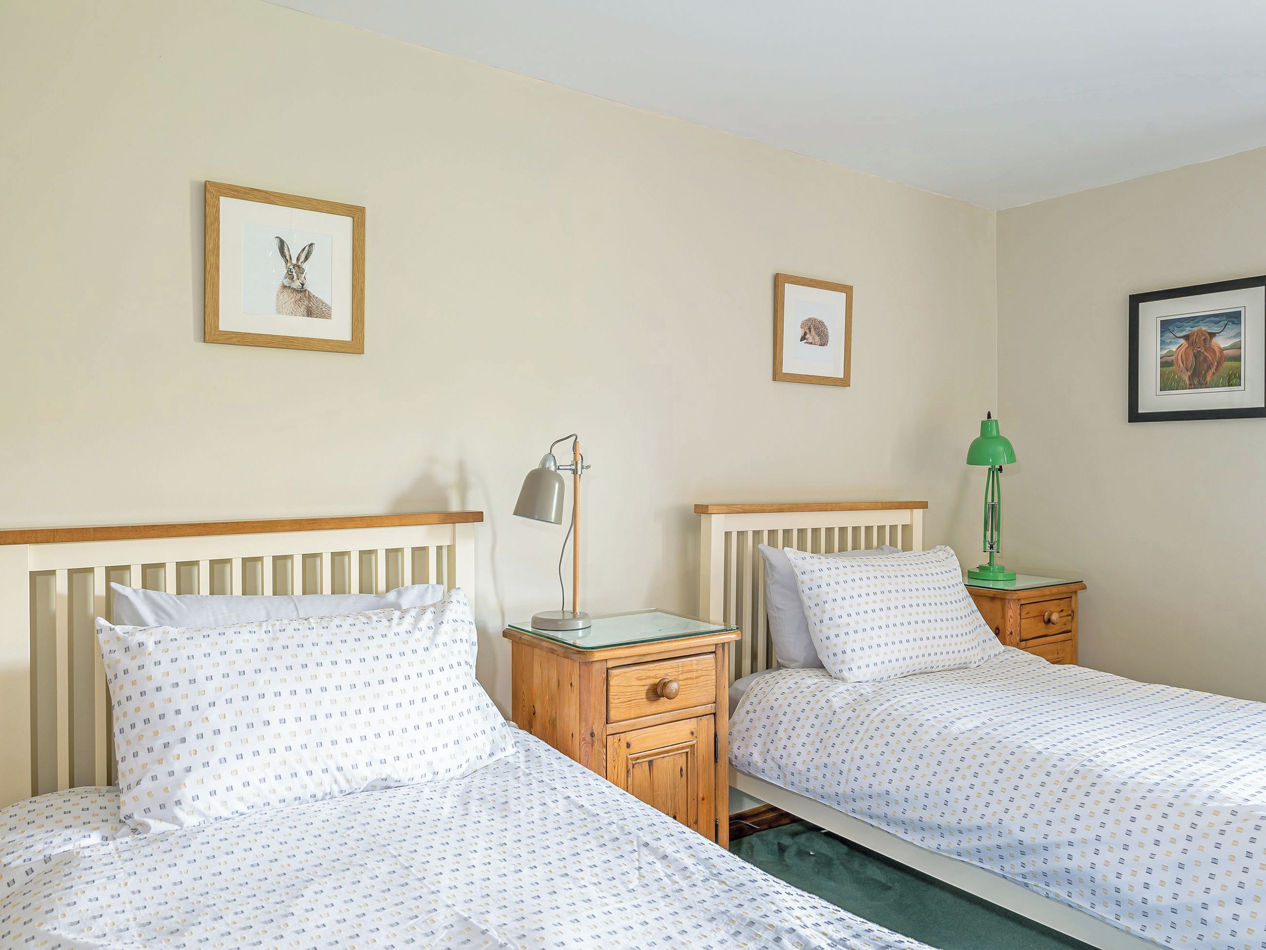 A room with two single beds, wooden headboards, bedside tables with lamps, and framed pictures on the walls.