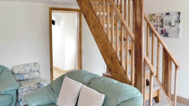 A living room with a green sofa, a wooden staircase, and a painting on the wall. - Deuglawdd Cottage - HW7788 (Aberdaron, near Pwllheli)