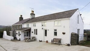 A white two-story house with a stone facade, a front porch, a bench, and a picnic table. - Deuglawdd Cottage - HW7788 (Aberdaron, near Pwllheli)