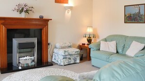 A living room with a fireplace, a sofa, a lamp, and a painting on the wall. - Deuglawdd Cottage - HW7788 (Aberdaron, near Pwllheli)