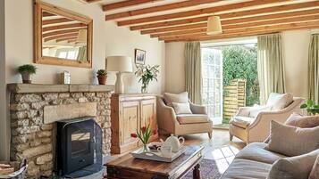 A cozy living room with a stone fireplace, wooden beams, comfortable seating, and a view of greenery outside.