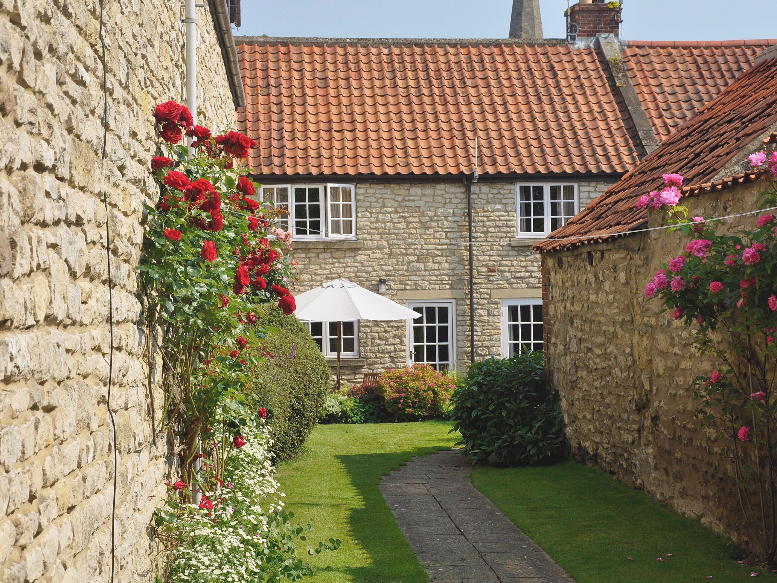 A stone building with a red-tiled roof, a paved pathway, and a garden with red and pink flowers.