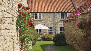 A stone building with a red-tiled roof, a paved pathway, and a garden with red and pink flowers.