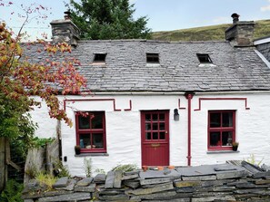 A white house with red windows and a red door, surrounded by stone walls and greenery.