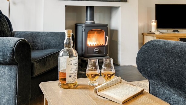 Un salón acogedor con chimenea, una botella de The Baive Signature de whisky escocés, dos vasos y un libro abierto sobre una mesa de madera.
