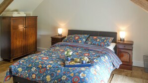 Une chambre avec un plafond en pente, un lit recouvert d’une couvre-lit à motifs floraux, deux tables de chevet avec des lampes et une armoire en bois.