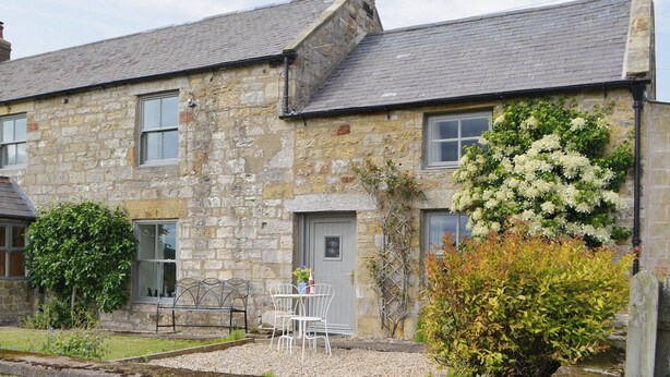 Une maison en pierre avec un toit recouvert de tuiles grises, une porte d’entrée et un petit jardin agrémenté d’un banc et d’une table.
