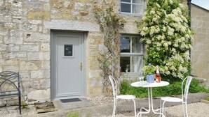 Un bâtiment en pierre avec une porte grise, une fenêtre et une table entourée de chaises à l’extérieur.