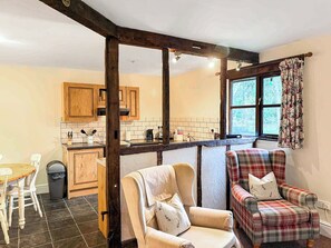 A kitchen with wooden cabinets, a dining table, and chairs. There is a window with curtains and a view of greenery outside.