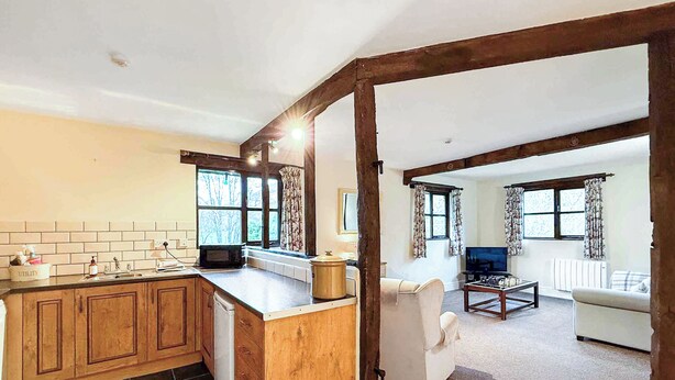 A kitchen with wooden cabinets, a microwave, and a sink. There is a dining area with a table and chairs, and a living room with a sofa and a television. - 1 bedroom accommodation in Newbridge-on-Wye, near Builth Wells (Newbridge-on-Wye, near Builth Wells)