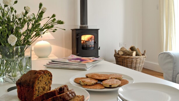 A cozy room with a wood-burning stove, a vase of flowers, a basket of bread, and a table set for breakfast. - A detached character cottage with beams, wood burner and enclosed garden. (Nanternis near New Quay)