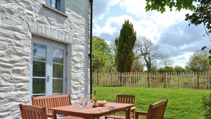 A stone building with a white door and windows, a wooden table with chairs, and a grassy yard with a wooden fence. - A detached character cottage with beams, wood burner and enclosed garden. (Nanternis near New Quay)