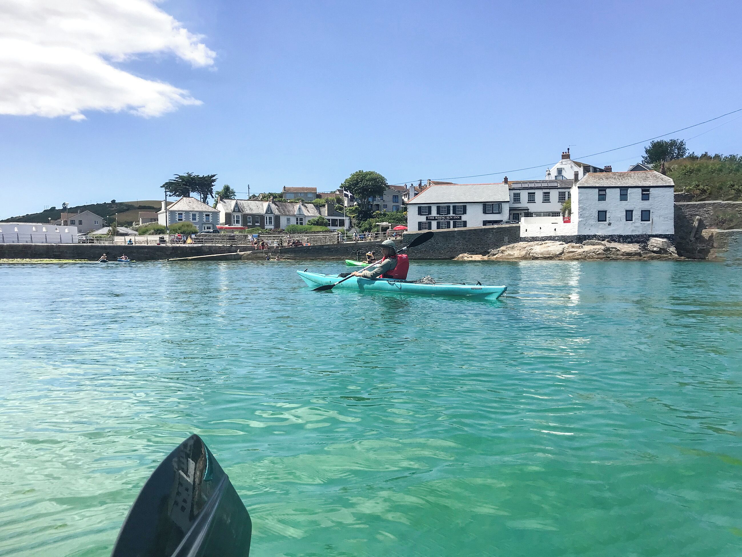 A clear waterfront view with a kayaker in a turquoise kayak, buildings in the background, and a stone wall along the shore.