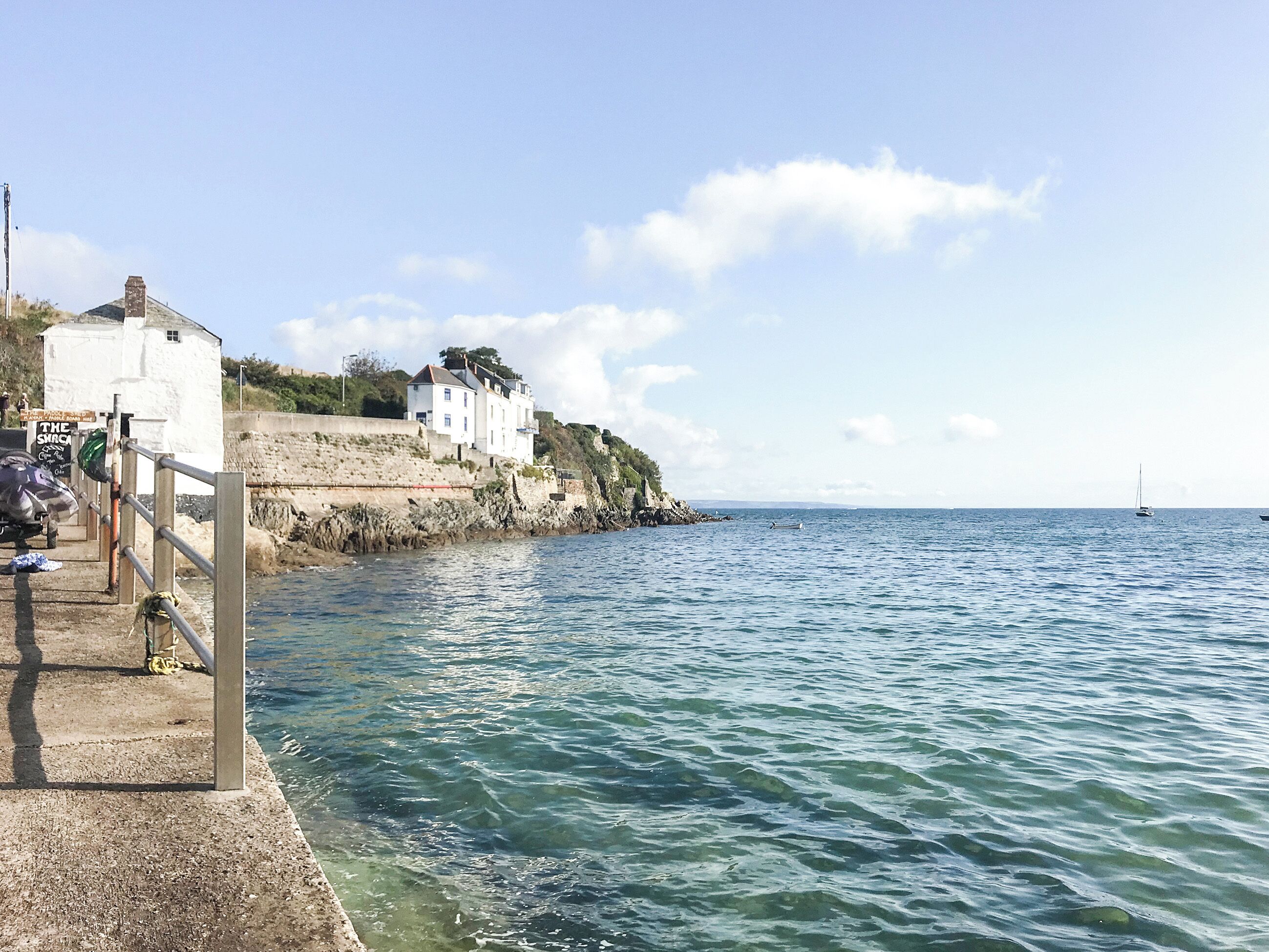 Un paysage côtier avec un bâtiment blanc perché sur une falaise surplombant la mer, une jetée en bois et un voilier à l’horizon.