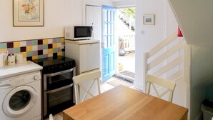 A compact kitchen with a washing machine, a microwave, and a stove. There is a wooden table and chairs, and a blue door leading outside.