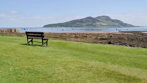 A grassy area with a bench facing a rocky beach and a body of water with boats.