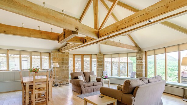 A spacious living room with wooden beams, a stone fireplace, and large windows offering a view of the outdoors. - 2 bedroom accommodation in Swainby, near Stokesley (Swainby, near Stokesley)