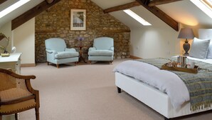 Une chambre avec un mur en pierre, deux fauteuils bleu clair, une chaise en bois, un lit recouvert d’une couverture à motifs, une table de chevet avec une lampe et une petite table sur laquelle est posé un vase.