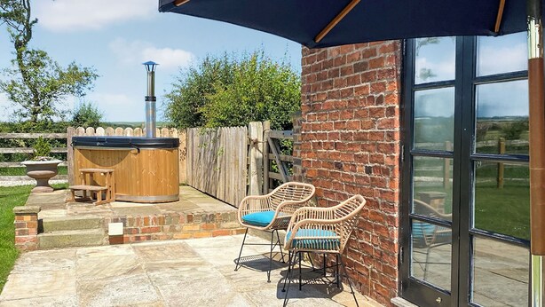 Une terrasse avec un mur en briques, une table en bois, deux chaises en osier et un grand parasol.