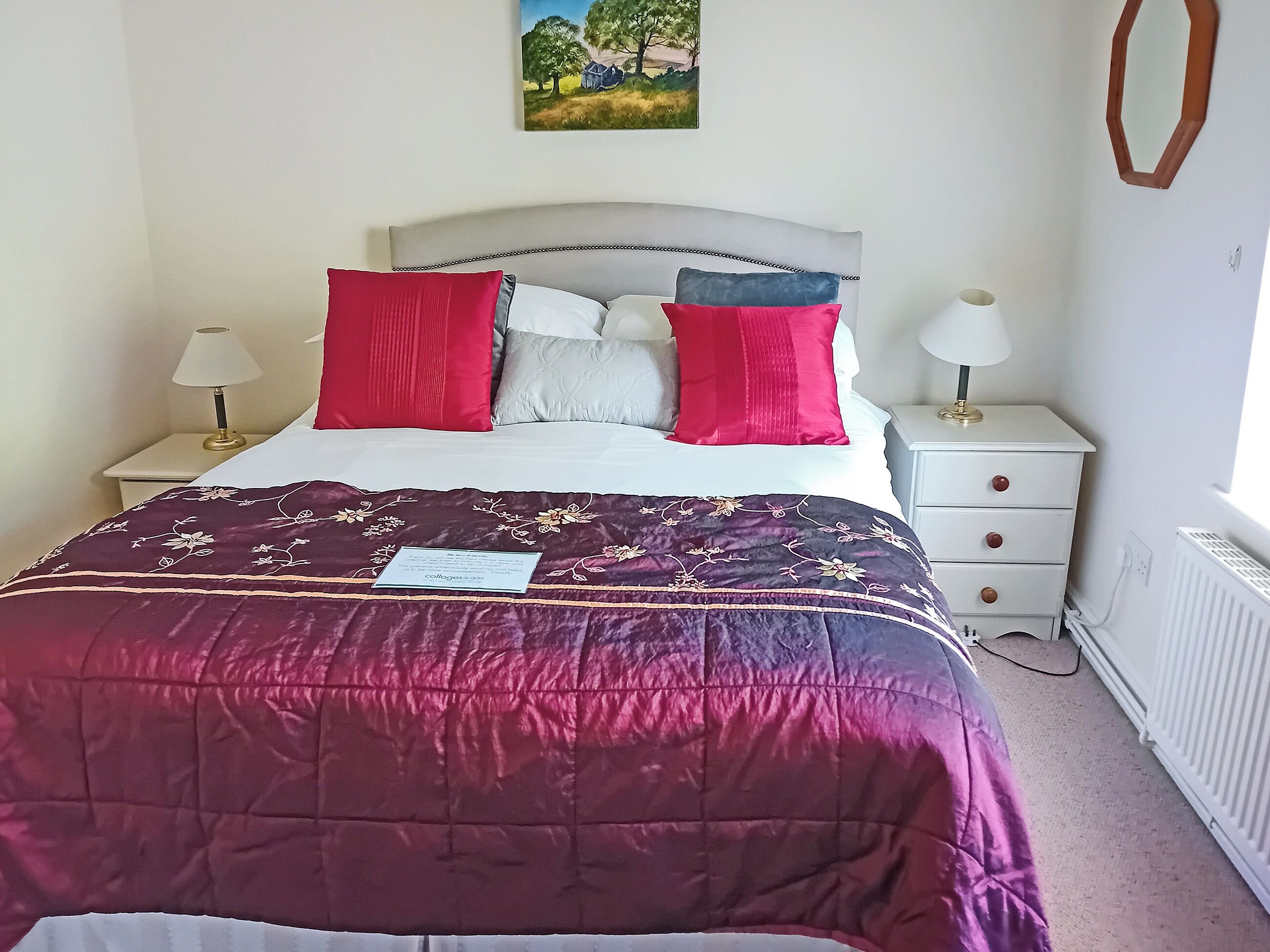 Un lit avec une tête de lit, une table de chevet avec une lampe, un radiateur et un miroir fixé au mur.