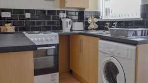 A modern kitchen with a stove, oven, and washing machine, wooden cabinets, black and white tiles, and a window with blinds. - 1 bedroom accommodation in Towyn, near Rhyl (Towyn, near Rhyl)