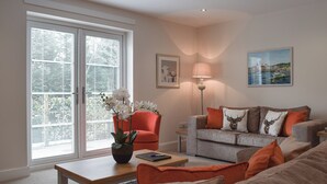 Una sala de estar con un sofá gris, cojines naranjas, una mesa de centro de madera, una lámpara de pie y una ventana con persianas.