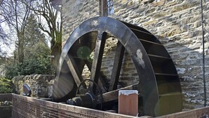 Una gran estructura de noria con armazón metálico y base de madera, ubicada frente a un muro de piedra.