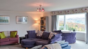 A living room with a sectional sofa, a blue armchair, and a bookshelf. There is a window with a view of hills.