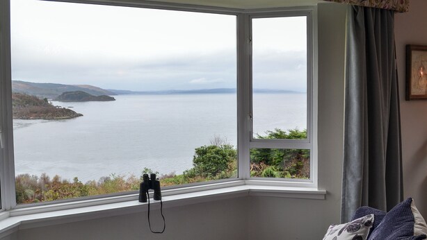 A room with a bay window overlooking a body of water and hills.