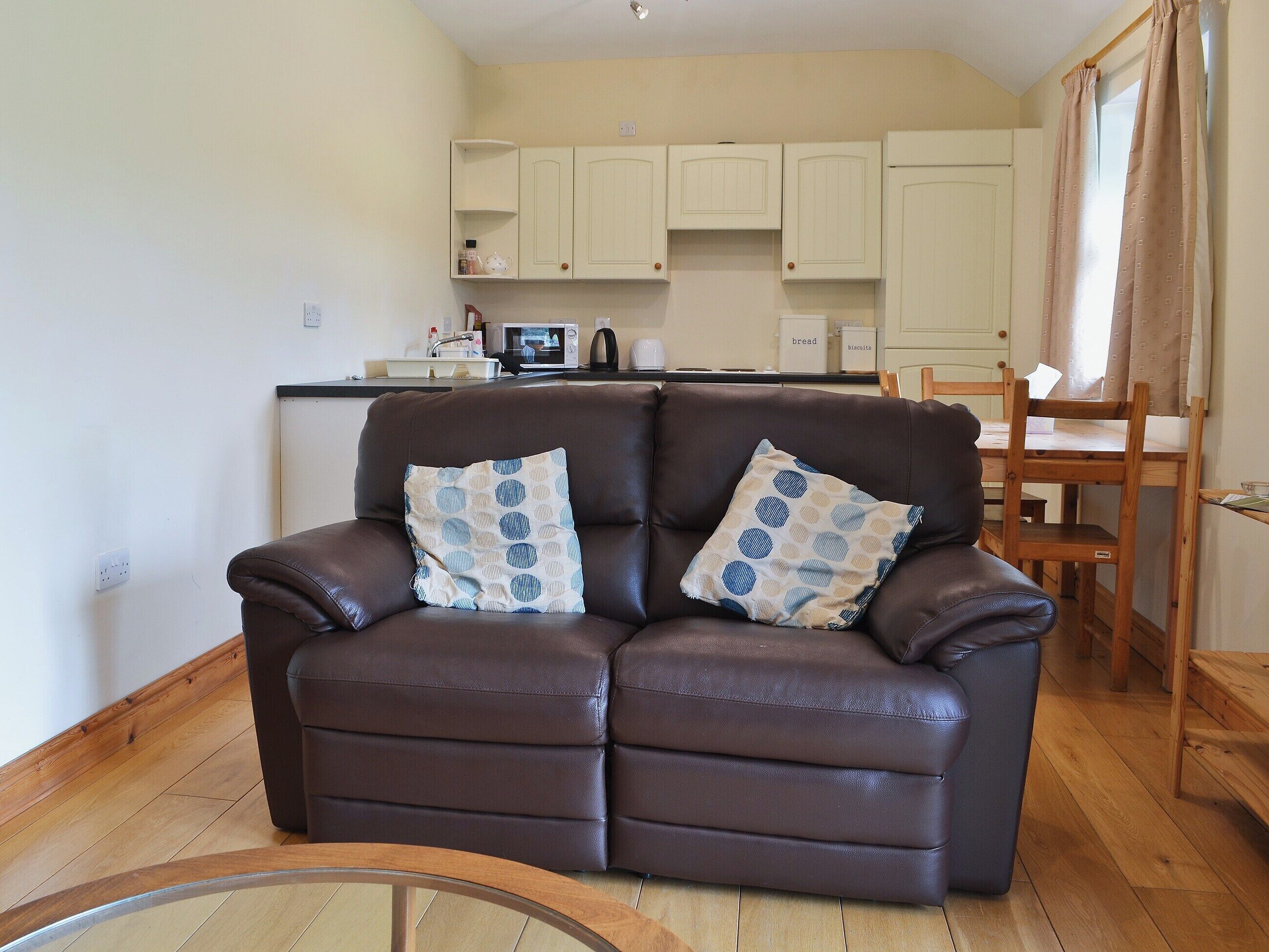 A brown leather sofa with two patterned cushions in a living room with wooden flooring. A kitchen with white cabinets and a dining area with a wooden table and chairs is visible in the background.