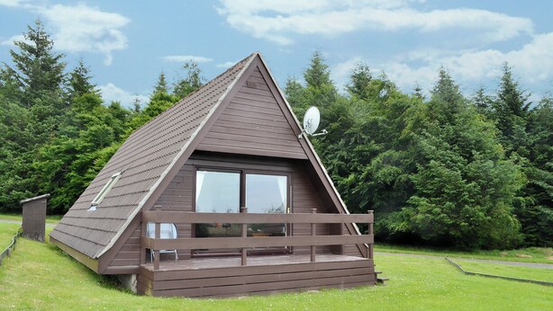 Eine kleine, dreieckige Holzhütte mit einem Balkon, umgeben von einer Wiese und dichtem Wald.