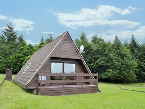 A small, triangular wooden cabin with a balcony, surrounded by a grassy area and dense forest.
