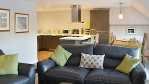 Una sala de estar moderna con un sofá de cuero negro, una mesa de comedor con sillas y una zona de cocina al fondo.