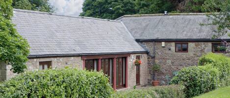 A stone building with a sloped roof, surrounded by lush greenery and a well-maintained lawn.