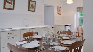 A dining area with a wooden table set for four, white plates, clear glasses, and a white candle. There are two framed pictures on the wall, a window with a view of greenery, and a ceiling light.