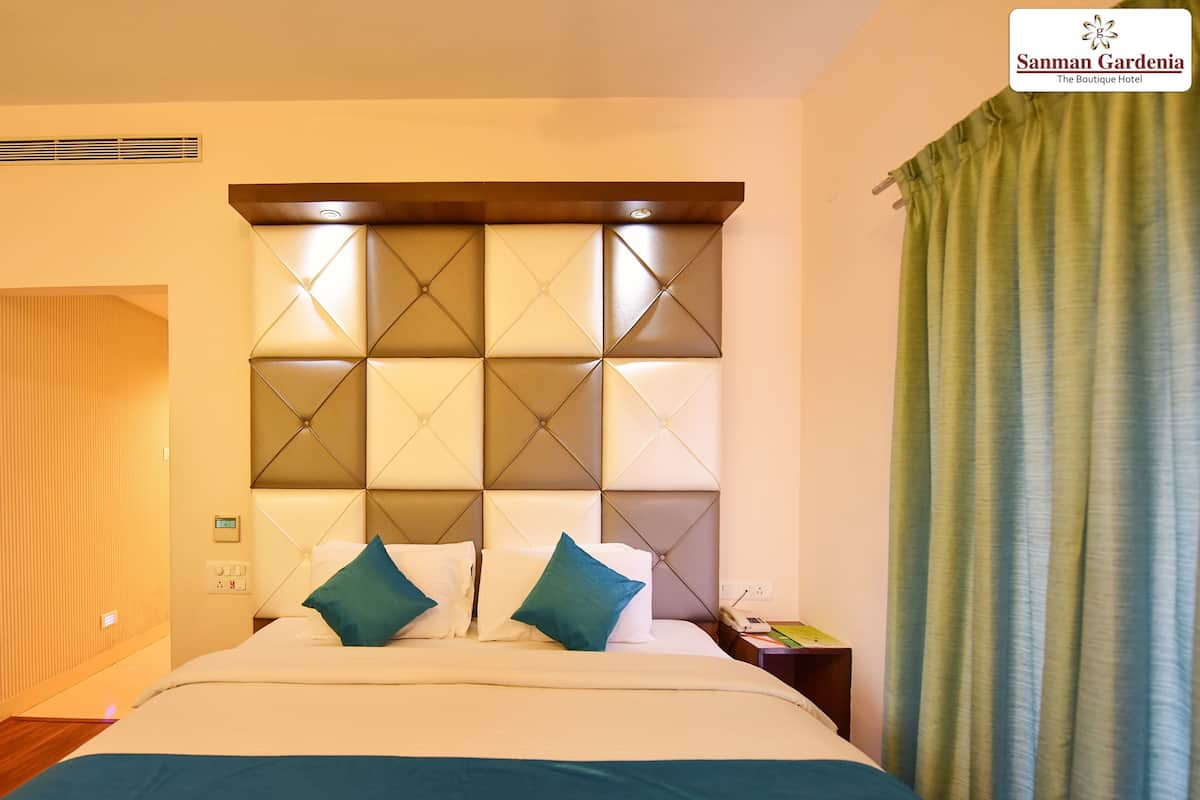 Una habitació d'hotel amb un capçal de llit amb borla, un llit amb roba de llit blanca i turquesa, una tauleta de nit amb una làmpada i una cortina verda.