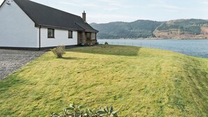 Une maison à l’extérieur blanc et au toit foncé, située au bord d’un lac, avec une pelouse verdoyante et un petit jardin.