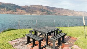 Une table de pique-nique avec des bancs, située au bord d’un lac, entourée d’une clôture et d’une zone herbeuse.